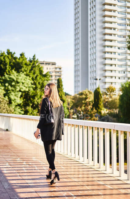 Rückenansicht einer positiven jungen Unternehmerin in schwarzem Mantel mit schwarzer Tasche beim Gang über die Brücke — Stockfoto
