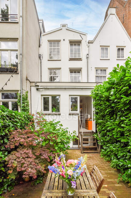 Двор современного жилого дома с деревянной садовой мебелью, украшенный букетом летних цветов среди яркой зелени в дневное время — стоковое фото