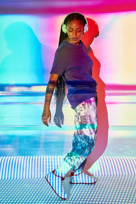 Vista lateral de la moda joven dominicana milenaria femenina con largas trenzas Afro de pie en el suelo y mirando hacia abajo mientras escucha música en los auriculares en la habitación con iluminación geométrica de colores - foto de stock