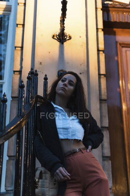 From below of serious young female in stylish outfit leaning on fence near entrance of building while looking away — Stock Photo