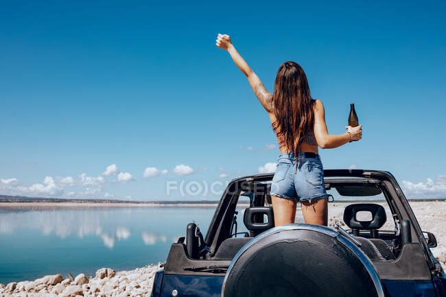 Rückansicht einer aufgeregten Frau im Sommeroutfit, die eine Hand Bier hebt, während sie auf dem Dach eines Safariwagens am Ufer des Flusses steht — Stockfoto
