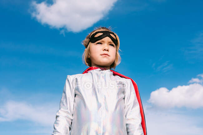 Знизу симпатична дитина в маскарадному костюмі супергероя і масці для очей, що стоїть на тлі блакитного неба і дивиться в сторону — стокове фото