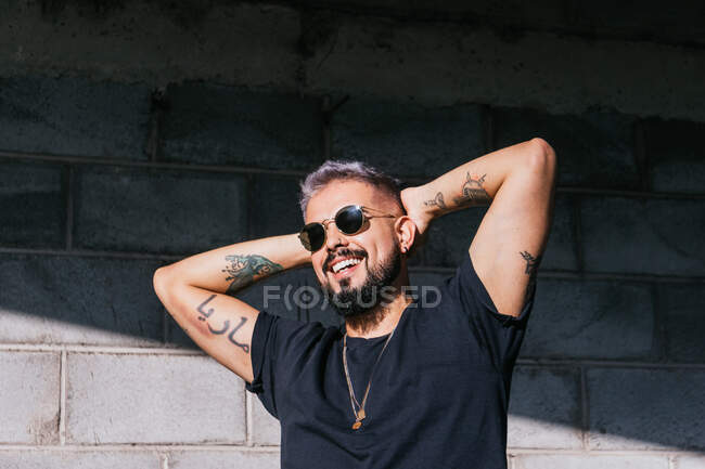 Hombre elegante sonriente con tatuajes en camiseta negra y gafas de sol de pie con los brazos levantados y mirando hacia otro lado contra la pared del edificio en el día soleado - foto de stock
