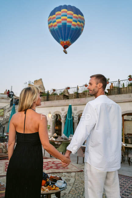 Back view of couple standing on the street cafe holding hands looking at each other while watching hot air balloon flight — Stock Photo