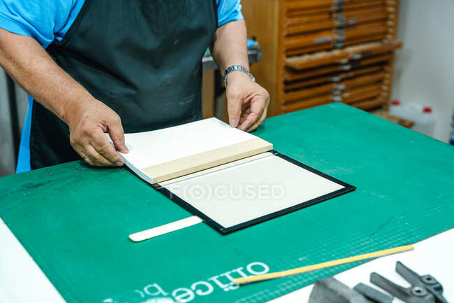 De cima de cultura anônima de meia-idade artesão masculino em avental fazendo ligação para livro de pé à mesa na oficina de impressão — Fotografia de Stock