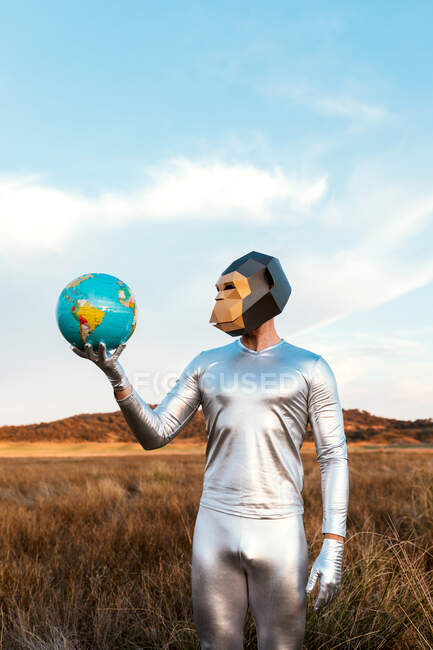 Chico anónimo en traje de látex plateado con máscara de mono geométrico mirando hacia otro lado y sosteniendo globo en la naturaleza - foto de stock