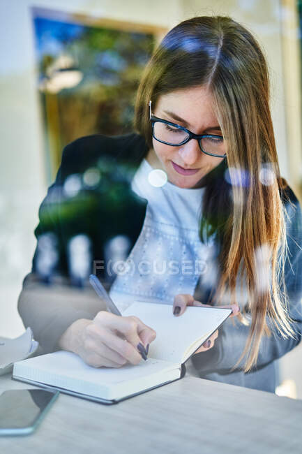 Durch das Glas einer konzentrierten Unternehmerin, die mit Smartphone am Tisch sitzt und Notizen zur Arbeit in den Notizblock schreibt — Stockfoto