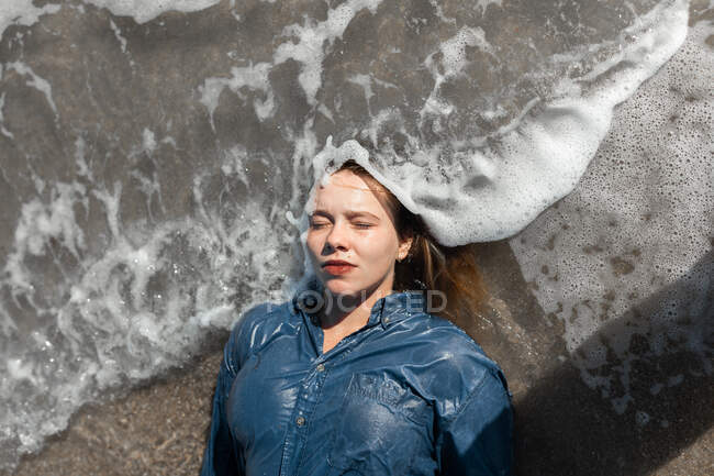 From above of calm female in wet shirt lying with closed eyes on sandy beach near waving sea on sunny summer day — Stock Photo
