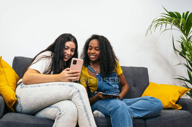 Young diverse female friends in casual clothes smiling while sitting on sofa and taking selfie in living room at home — Stock Photo