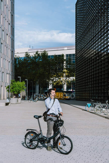 Full body of male worker in formal wear standing with bicycle and having conversation on mobile phone in downtown — Stock Photo