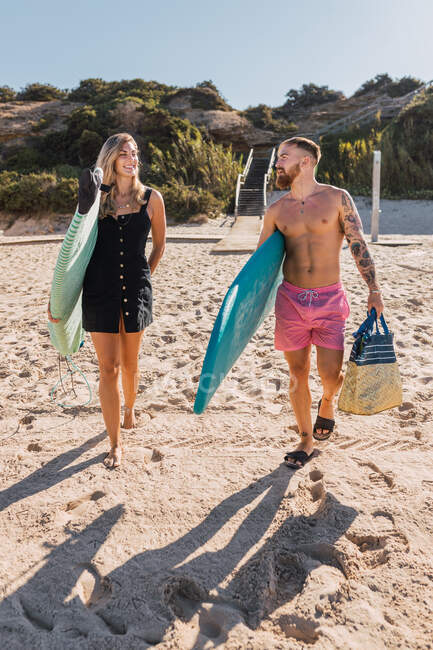 Повне тіло щасливої спортивної пари з дошками для серфінгу, що прогулюються разом на піщаному пляжі перед тренуванням у морі — стокове фото