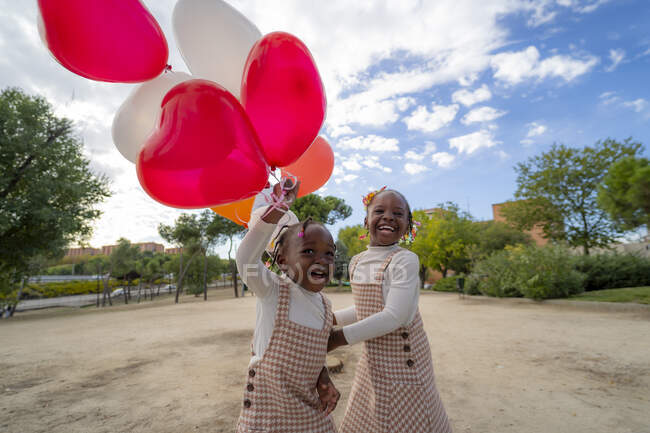 Hermanitas afroamericanas felices con vestidos similares de pie con globos de colores en las manos sobre hierba verde en el parque a la luz del día - foto de stock