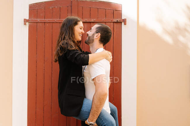 Вид збоку романтичного молодого етнічного бородатого чоловіка в повсякденному одязі, що носить щасливу дівчину з закритими очима біля дверей на вулиці в сонячний день — стокове фото