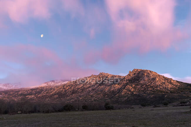 Impresionante paisaje de cordillera rocosa y valle con árboles verdes bajo el cielo rosado del atardecer con nubes y luna en el Parque Nacional Sierra de Guadarrama en España - foto de stock