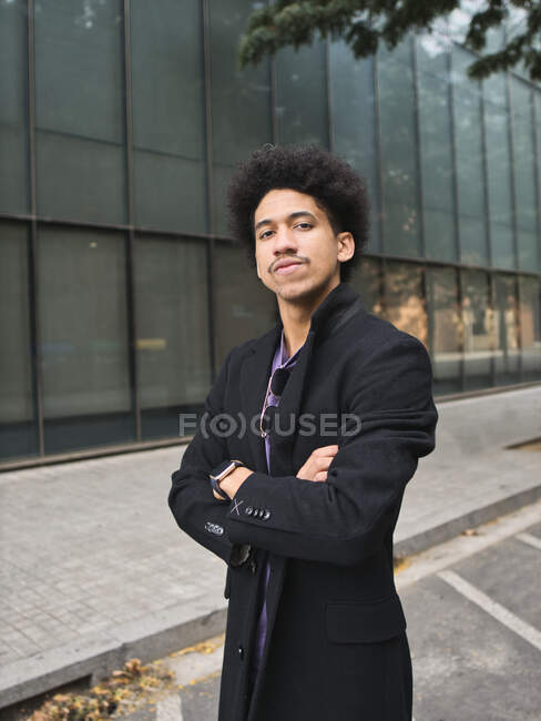 Empresario masculino étnico joven con cabello afro oscuro ajustando el traje elegante y mirando a la cámara mientras está parado en la calle cerca del edificio moderno - foto de stock