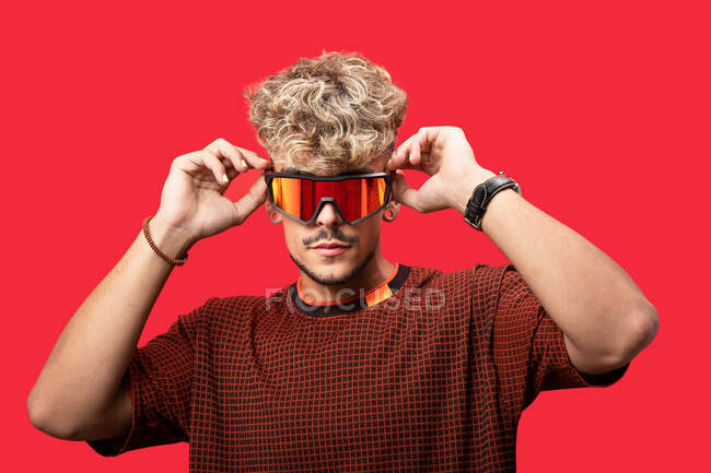 Серйозний молодий чоловік з кучерявим волоссям в модних сонцезахисних окулярах, що стоять на червоному тлі і дивляться на камеру — стокове фото