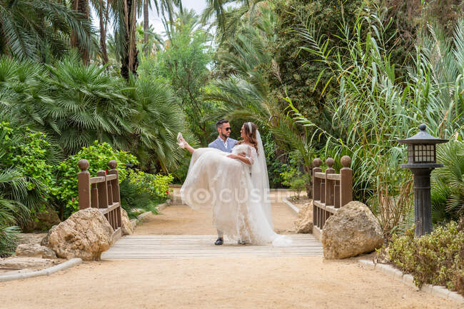 Ganzkörper-Bräutigam hält Braut im weißen Kleid in den Händen, während sie während des Hochzeitsurlaubs neben grünen tropischen Bäumen steht — Stockfoto