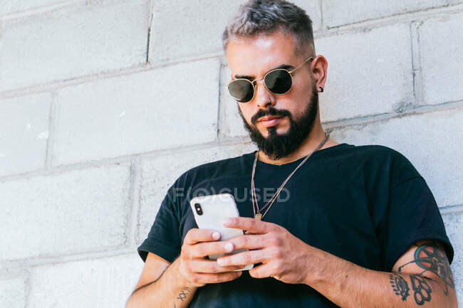 Happy bearded guy with tattoos in black t shirt and sunglasses standing near wall of building and using a smartphone in daylight — Stock Photo