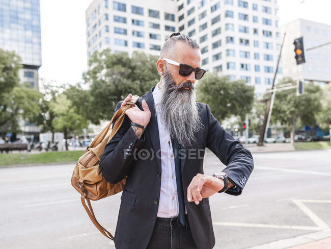 Confident male hipster in sunglasses and formal outfit with bag checking time on wristwatch while standing near road on street with buildings — Stock Photo