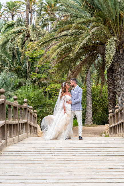 Супружеская пара в свадебных нарядах, стоящая на деревянном пешеходном мосту с перилами, обнявшись и глядя друг на друга возле зеленых пальм и растений в саду в летний день — стоковое фото