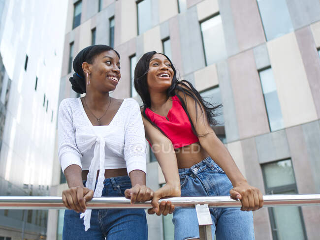 Von unten entzückte afroamerikanische Freundinnen in stylischen Outfits, die wegschauen, während sie sich an ein Metallgeländer in der Nähe eines modernen Gebäudes in der Stadt lehnen — Stockfoto