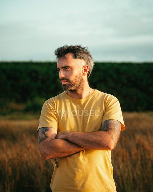 Hombre seguro con tatuajes en camiseta casual de pie en prado herboso con brazos cruzados en la naturaleza en el día de verano - foto de stock