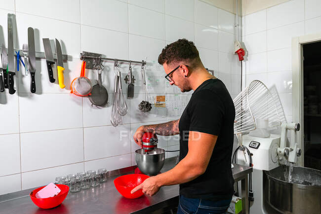 Вид сбоку на молодого татуированного мужчины-кондитера в повседневной одежде и очках, взвешивающего муку в электронном масштабе, стоящего за столом с миксером на кухне — стоковое фото