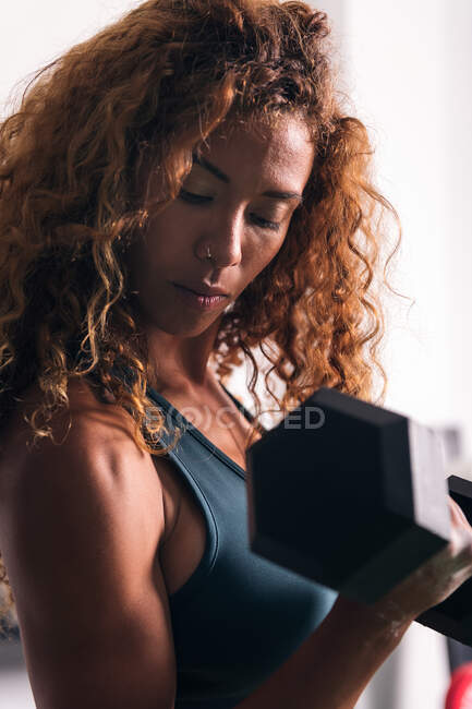 Vista lateral de una deportista fuerte con cabello rizado haciendo ejercicio en bíceps con mancuerna durante el entrenamiento en el gimnasio - foto de stock