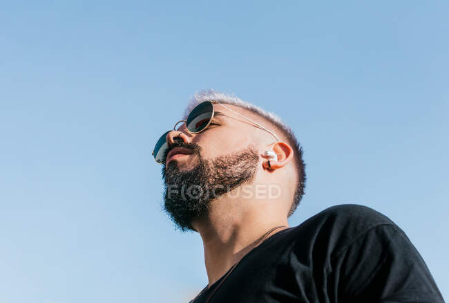 Знизу спокійного чоловіка з чорною бородою і вусами в сонцезахисних окулярах і повсякденному одязі, дивлячись на безхмарне блакитне небо на сонячному світлі — стокове фото