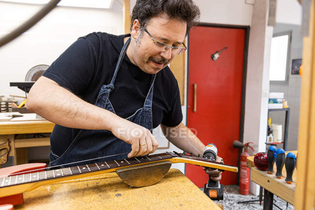 Concentrated adult man in apron standing near table with guitar while using drill to fix instrument near screwdrivers in bright room — Stock Photo