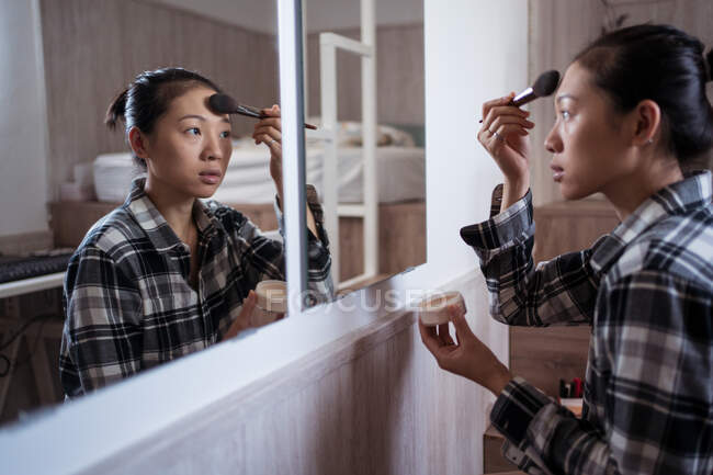 Vue latérale de la jeune femme asiatique en vêtements décontractés appliquant de la poudre tout en se tenant devant le miroir dans la chambre à coucher lumineuse — Photo de stock