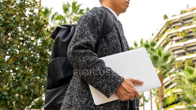 Side view of cropped unrecognizable male entrepreneur with rucksack and netbook looking away against modern urban buildings and palm trees — Stock Photo