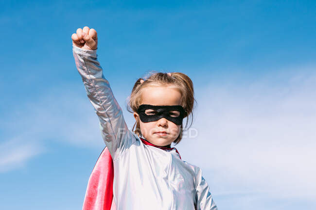 Desde abajo pequeña niña en traje de superhéroe levantando puños extendidos para mostrar poder mientras está de pie contra el cielo azul claro - foto de stock