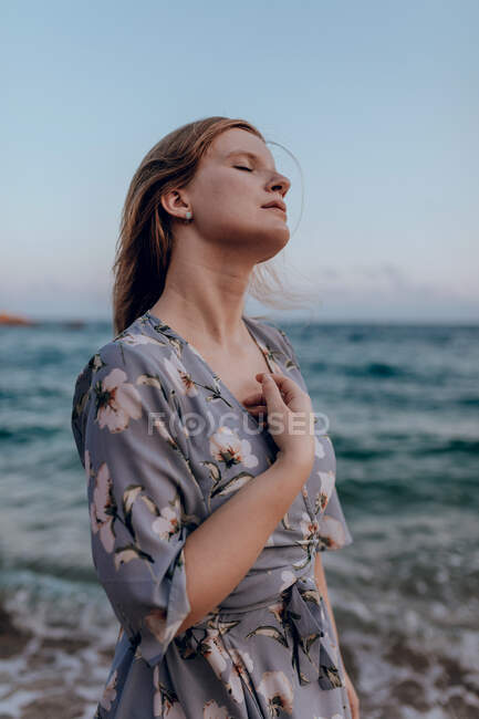 Entzückte Frau mit langen Haaren im trendigen Kleid steht mit geschlossenen Augen an der Küste im Sommerabend — Stockfoto