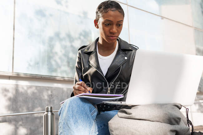 Femme afro-américaine concentrée dans une veste en cuir parcourant le netbook tout en prenant des notes dans un carnet dans la rue près du bâtiment en ville — Photo de stock