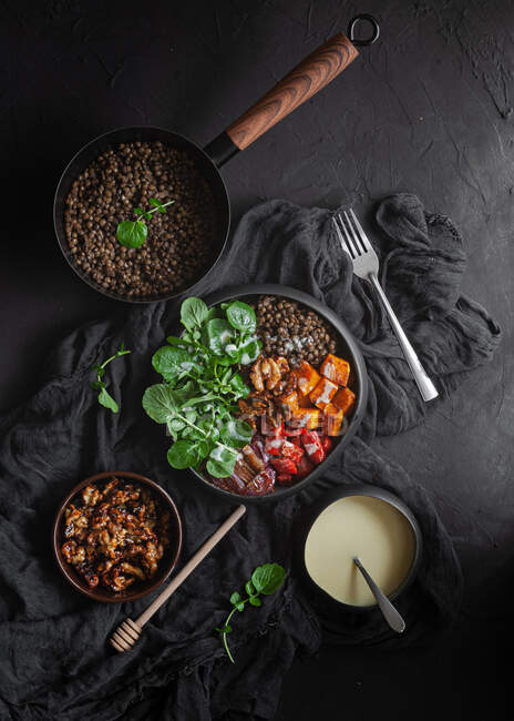 Vue du dessus de la casserole aux lentilles près de la salade avec citrouille et poivron décoré de feuilles de basilic vert sur fond noir près de la vinaigrette — Photo de stock