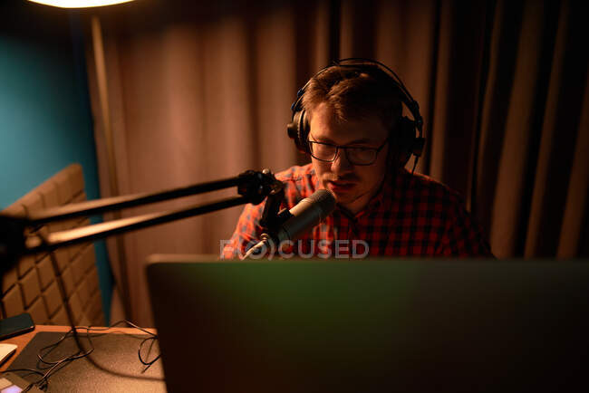 From above focused young male in checkered shirt and eyeglasses using computer and speaking in mic while recording podcast in dark studio — Stock Photo