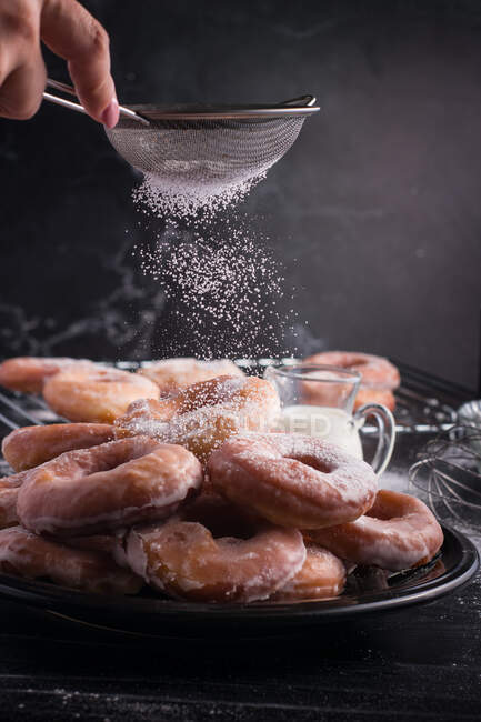 Crop anonymous chef with strainer sprinkling powdered sugar on heap of sweet fried doughnuts served on plate on gray background — Stock Photo