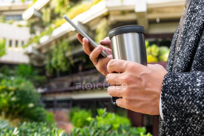 Side view of crop anonymous male with hot drink in tumbler text messaging on cellphone in town on blurred background — Stock Photo