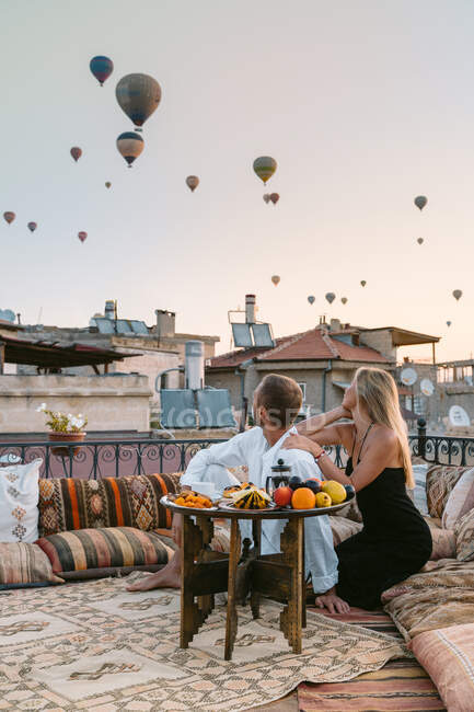 Anonymes Paar sitzt zusammen auf dem Fußboden im Dachcafé und betrachtet jede Menge fliegende Heißluftballons am Abend — Stockfoto
