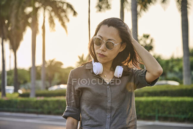 Serious Asian female with modern white headphones looking into distance while standing near road on street of town with green trees — Stock Photo