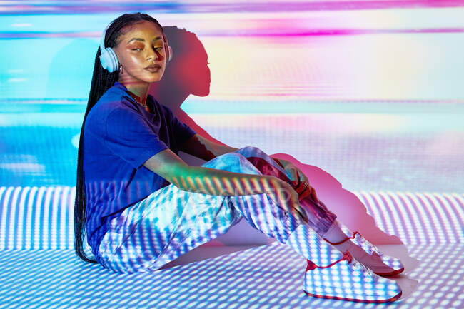 Vista lateral de la moda joven dominicana milenaria femenina con largas trenzas Afro sentado en el suelo y mirando hacia otro lado mientras escucha música en auriculares en la habitación con iluminación geométrica colorida - foto de stock