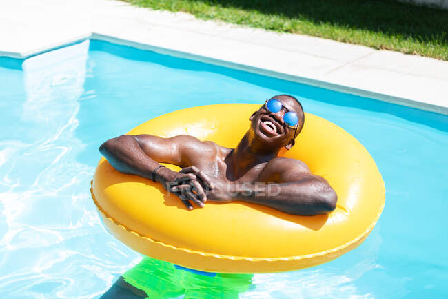 Calma hombre negro en pantalones cortos de natación y gafas de sol tomar el sol en el anillo inflable amarillo en la piscina con agua clara en el día de verano soleado - foto de stock