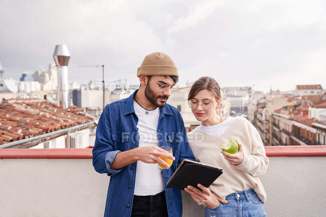 Молодий чоловік зі склянкою апельсинового соку, що стоїть поруч із сусідкою, їсть здорове зелене яблуко на даху та переглядає планшет разом — стокове фото