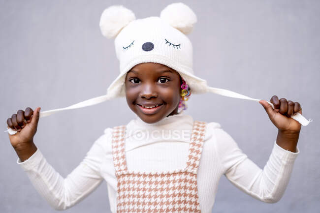 Cheerful African American little girl in stylish clothes and funny hat standing with raised arms and looking at camera against gray background — Stock Photo