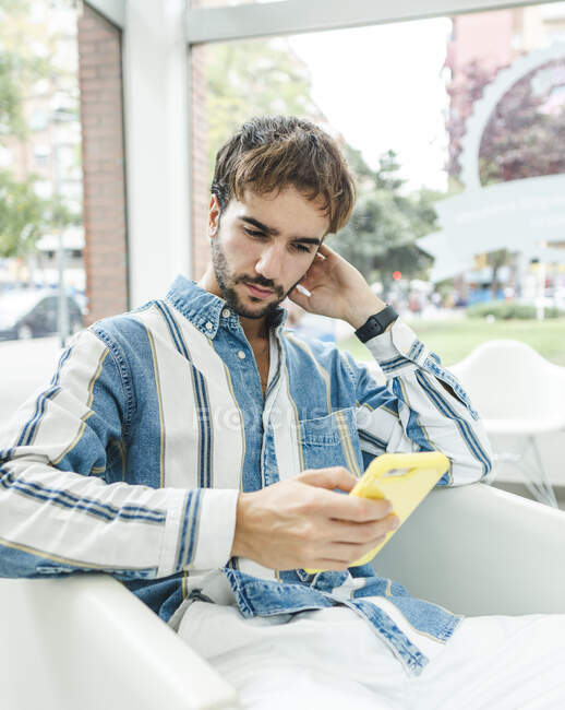 Homem barbudo concentrado em t-shirt listrada sentado em poltrona contra janela e smartphone de navegação — Fotografia de Stock