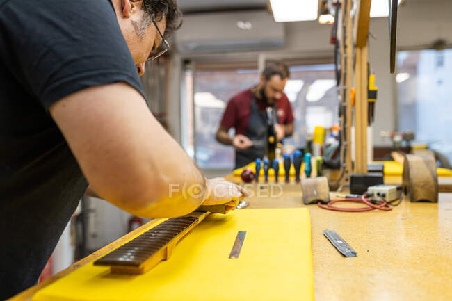 Focused guitar master standing at table and crowning frets on guitar neck during work with colleague in professional workshop — Stock Photo
