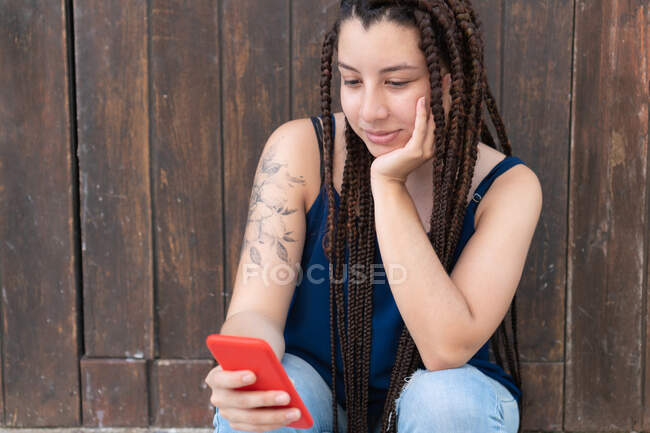 Konzentrierte hispanische Frau mit Tätowierung und langen geflochtenen Haaren SMS-Nachrichten auf dem Handy, während sie in der Nähe der hölzernen Mauer auf der Straße sitzt — Stockfoto