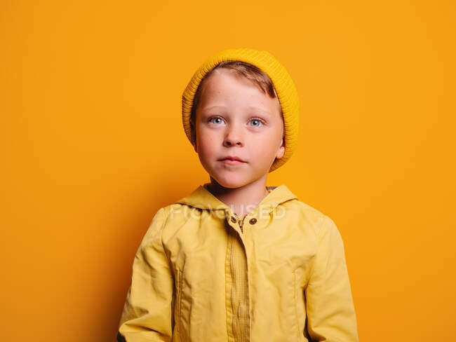 Niño poco emocional en impermeable de moda y gorro sombrero de pie mirando hacia otro lado contra el fondo amarillo en el estudio - foto de stock