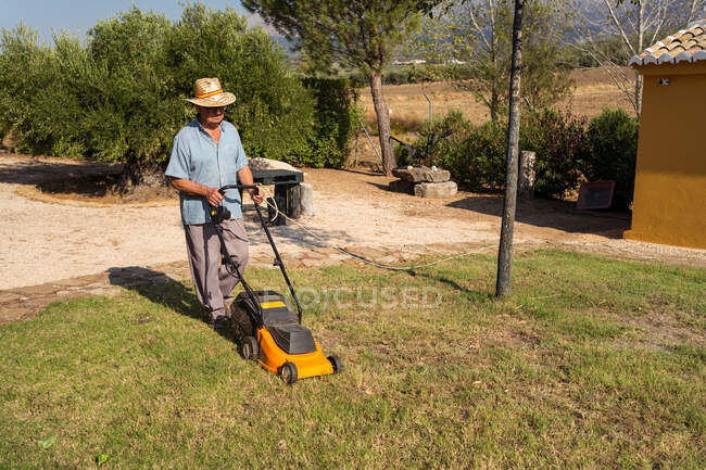 From above full body of unrecognizable male gardener in hat mowing grassy lawn near bushes and trees in summer — Stock Photo